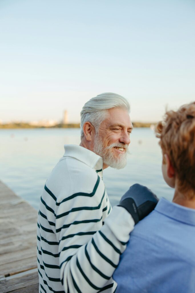 Smiling elderly man with white hair and boy in stripes sitting on a wooden dock by a lake.